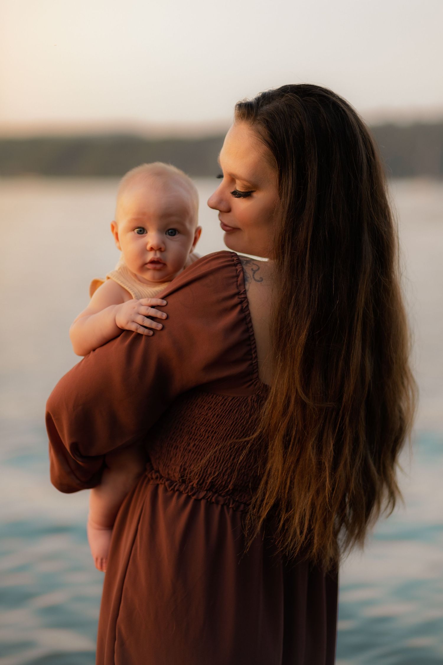 Person in brown dress holds baby against sunset lake backdrop.