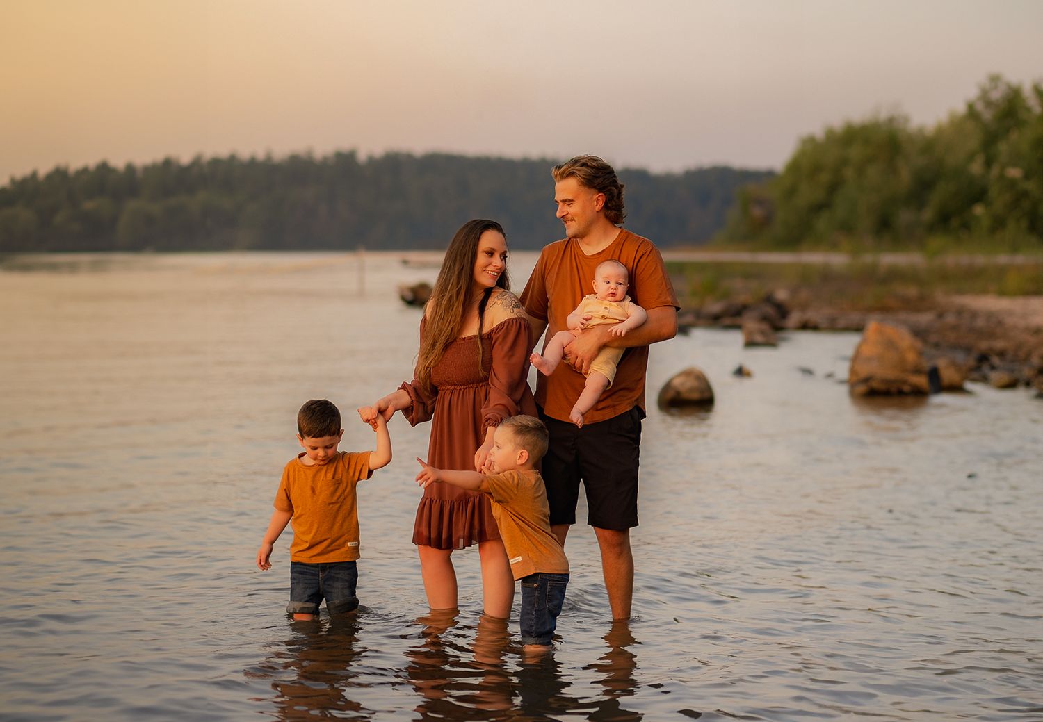 Family wades in shallow water at sunset near rocky shoreline.