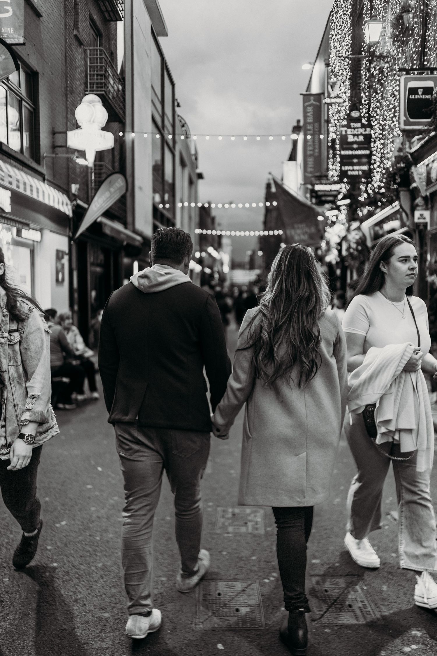 People walking through a bustling street with string lights and shops in black and white.