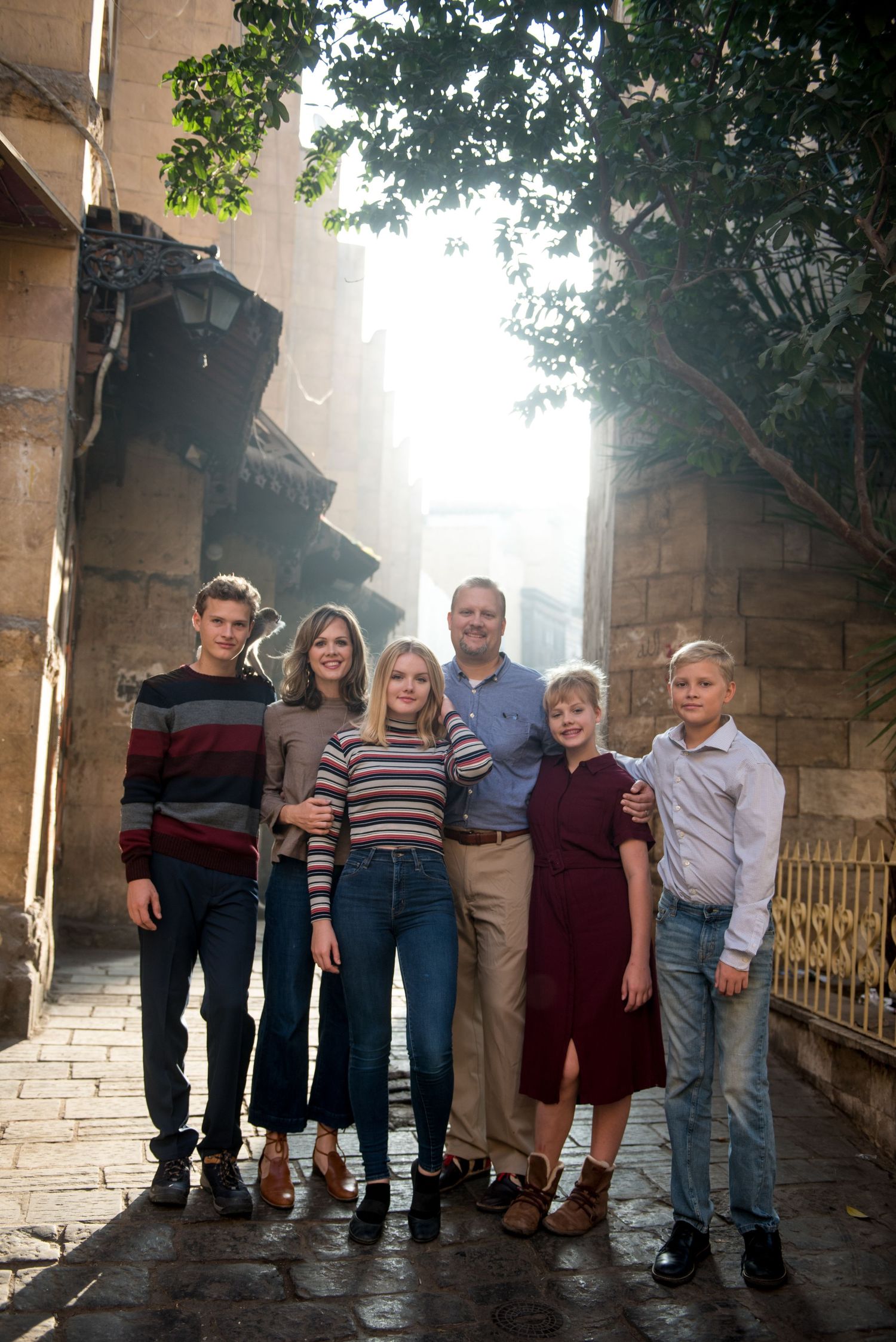 A group stands together on a stone path under an archway with trees and sunlight streaming through the background.