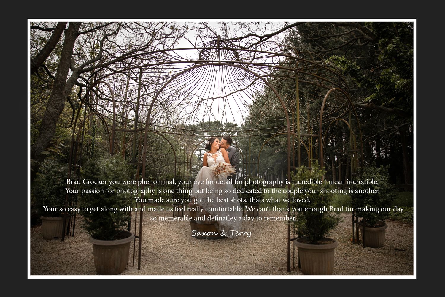 Couple stands under decorative iron archway in a garden setting.
