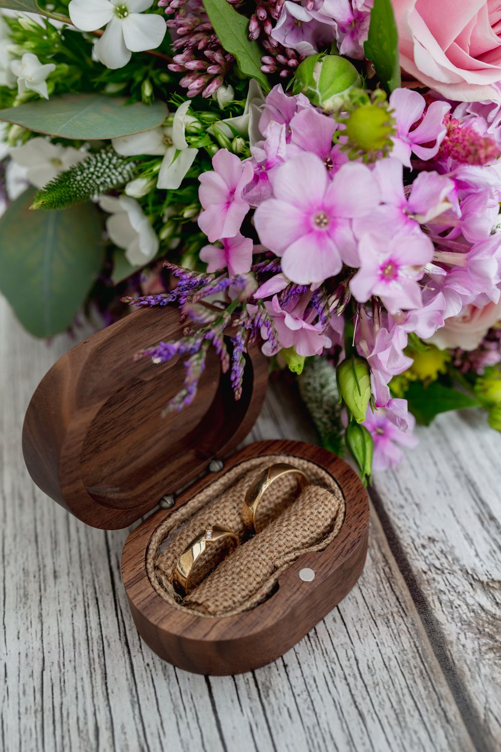 Wedding rings in wooden box with pink and purple floral arrangement on weathered wood surface.
