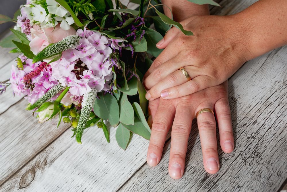 Wedding rings and pink flowers arranged on rustic wooden surface with hands showing.