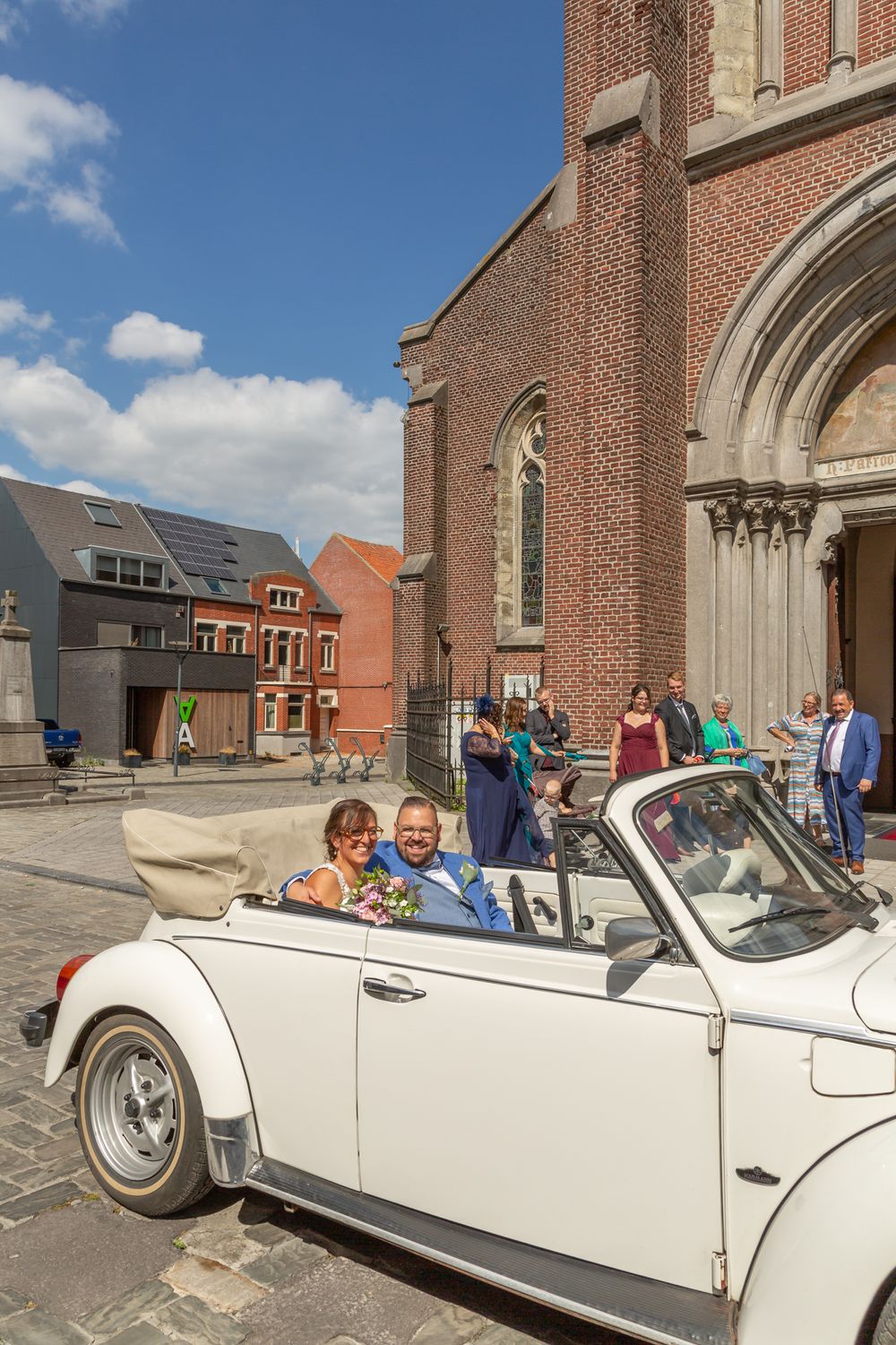 Classic white vintage convertible parked in front of a brick church with people gathered outside on a sunny day.
