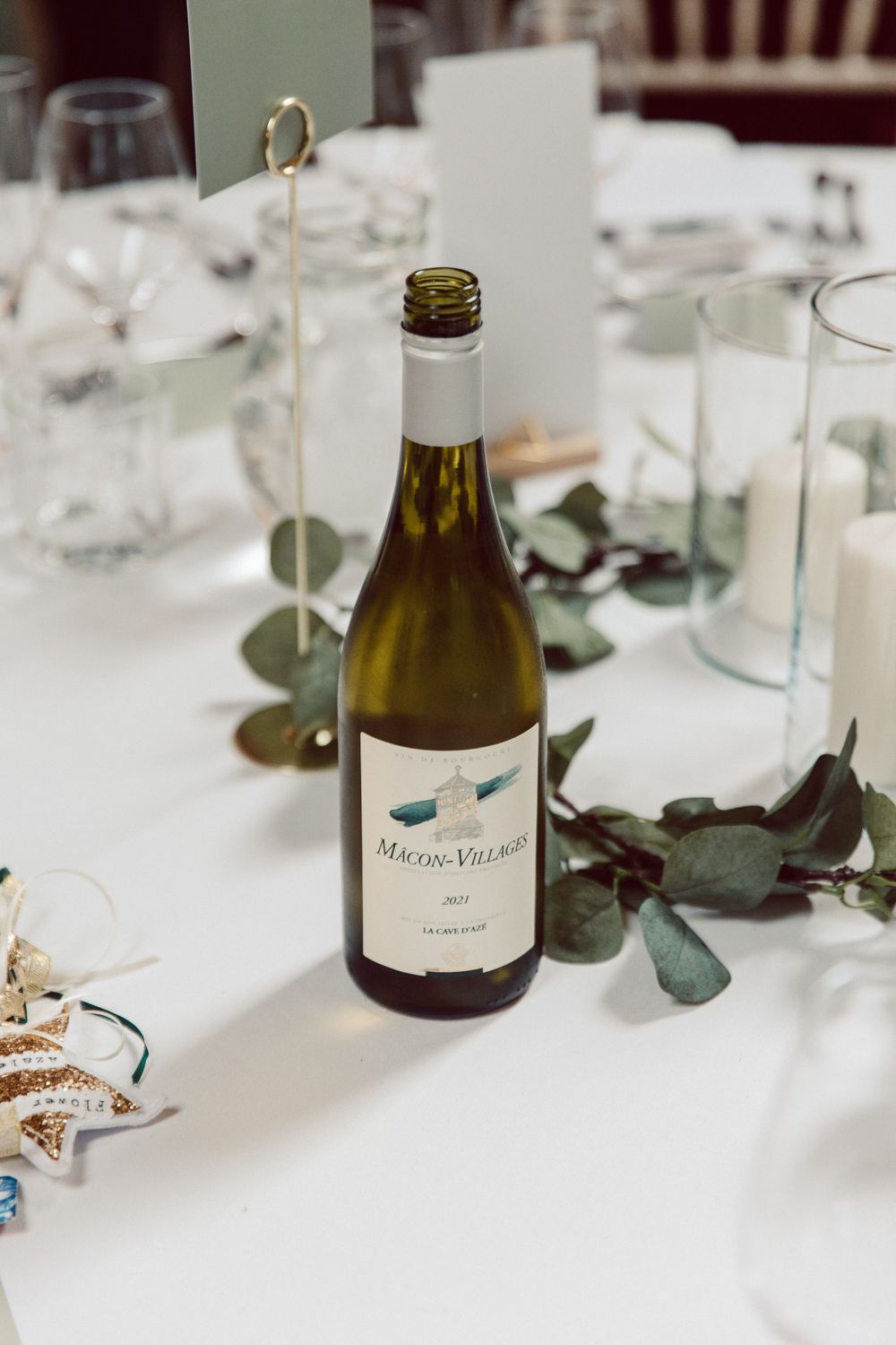 Wine bottle and table settings with eucalyptus leaves on white tablecloth at formal event.