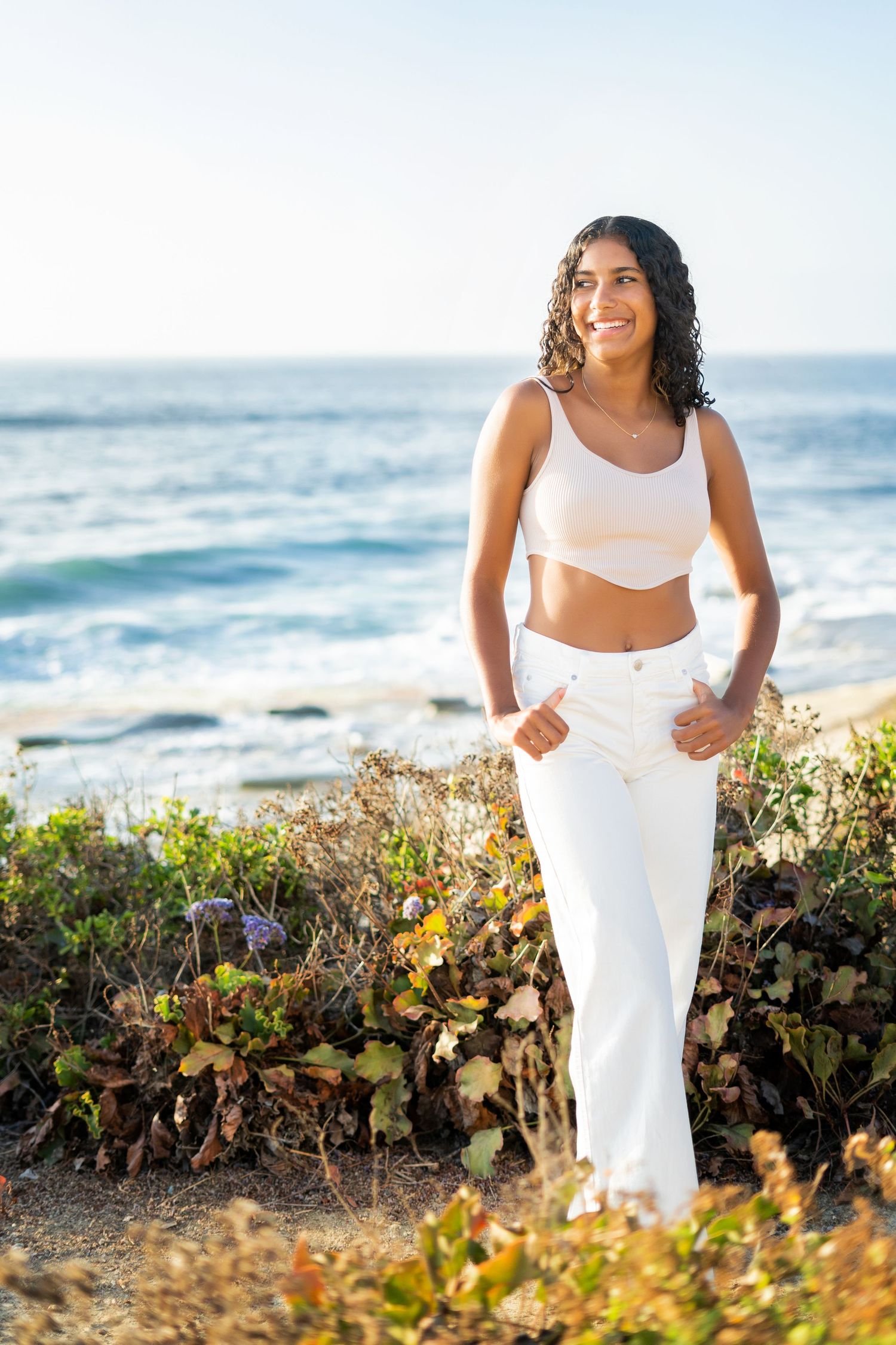 A high school senior in white workout attire posing by ocean with waves in background.