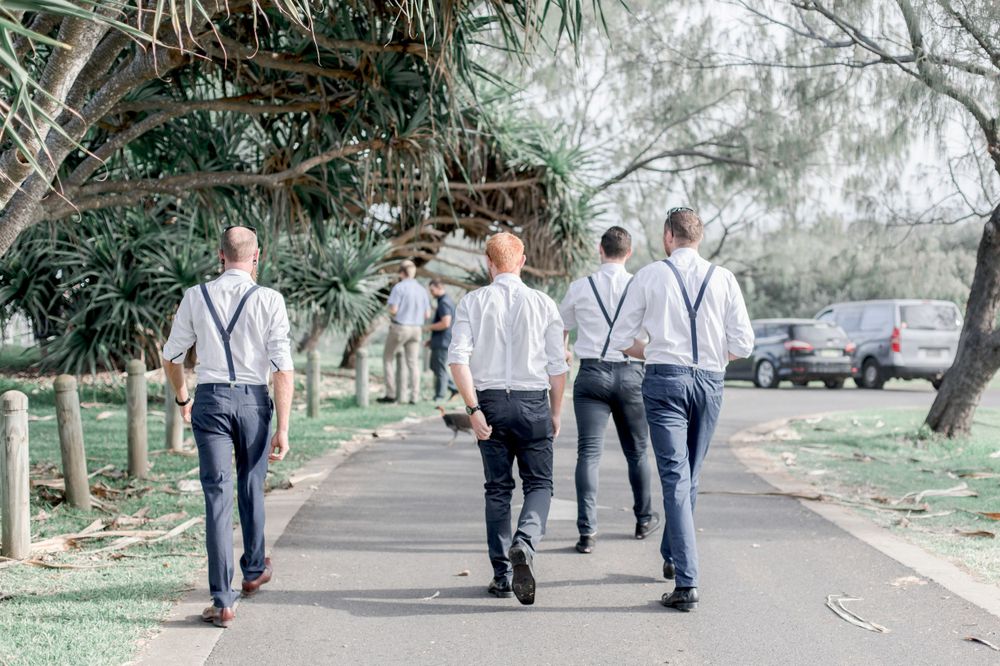 group of men in pants and suspenders walking down pathway to wedding, groom is scratching his bottom while turkey crosse