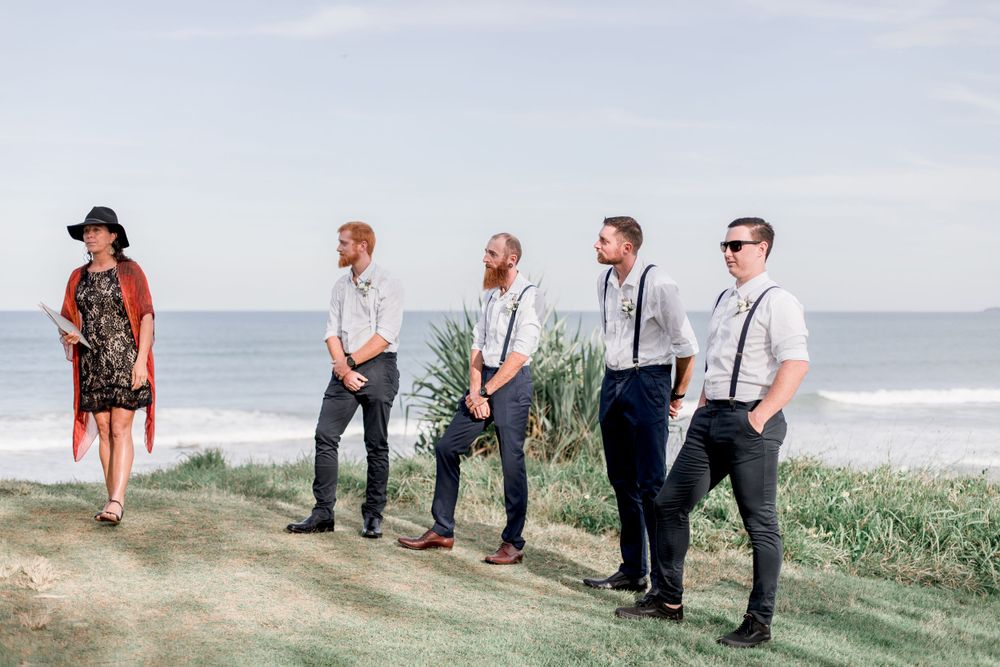 grooms party in suspenders and white shirts standing on cliff top waiting for bride with celebrant wearing hat smiling