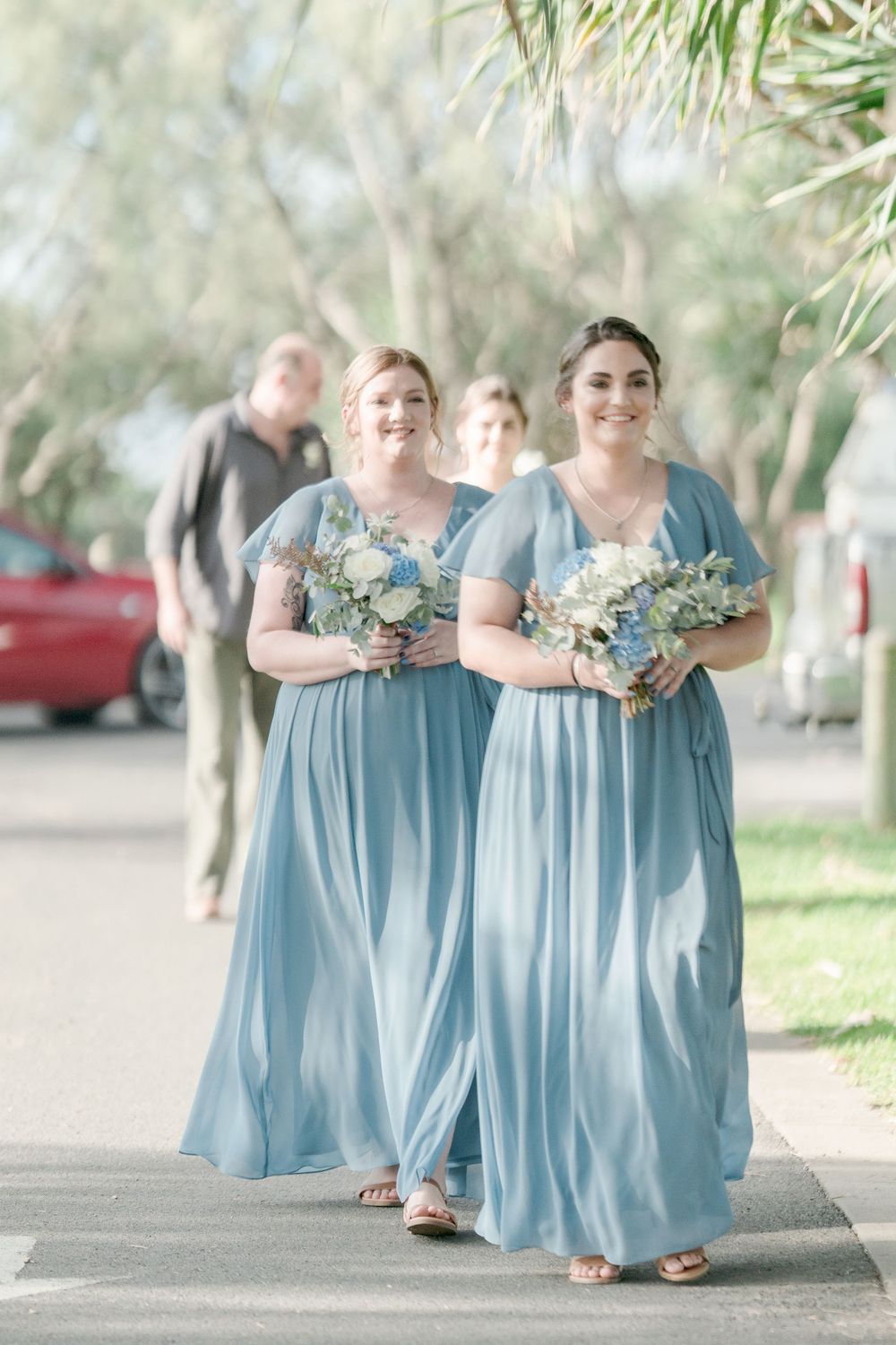 two women in long pale blue gowns carrying blue and white bouquets walking down road under trees