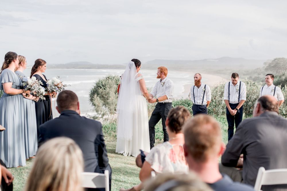 cliff top wedding with ocean view and bridesmaids wearing pale blue gowns and groomsmen in suspenders viewed over heads