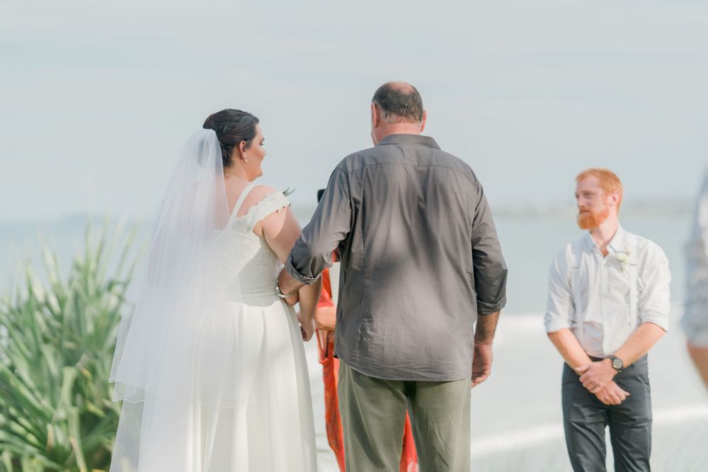 plus size brunette bride in veil and long gown being walked down aisle by father in grey shirt with redheaded groom wait