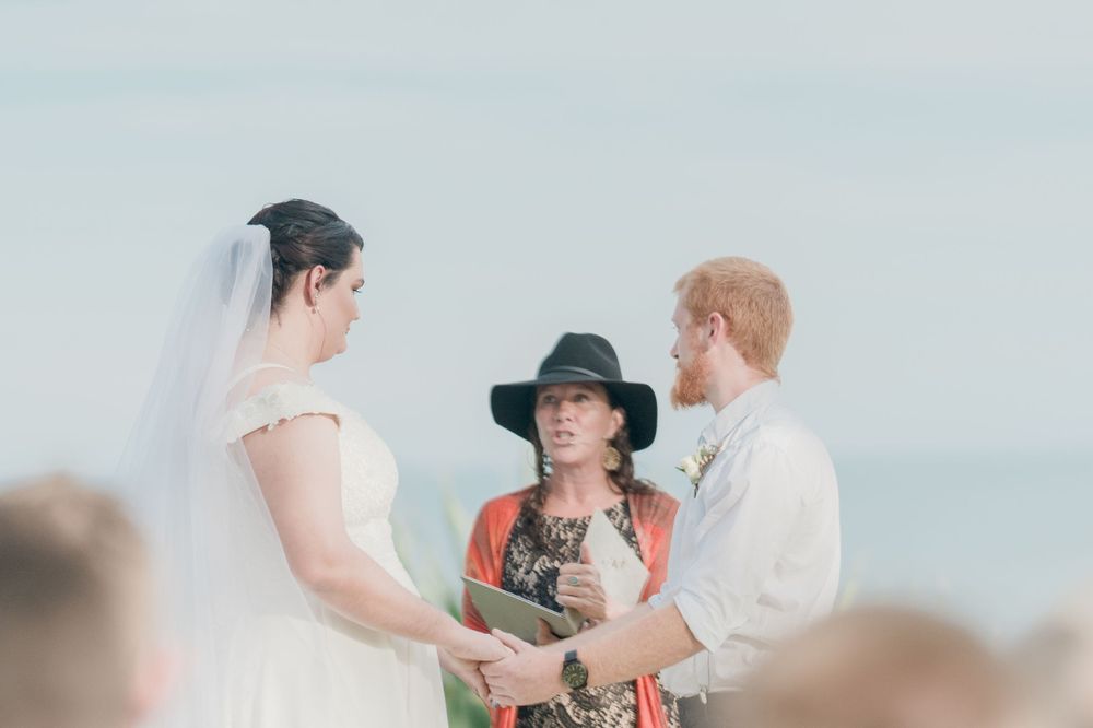 celebrant in red jacket and black hat performing wedding ceremony for plus size bride and redheaded groom overlooking oc