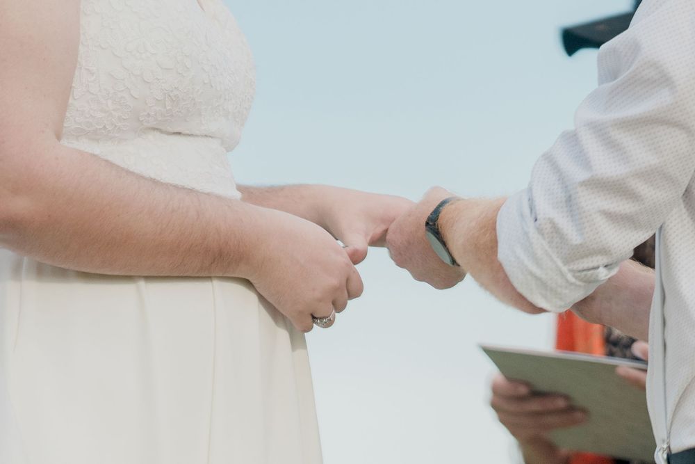 groom wearing black watch placing wedding ring on finger of plus size bride in sleeveless dress