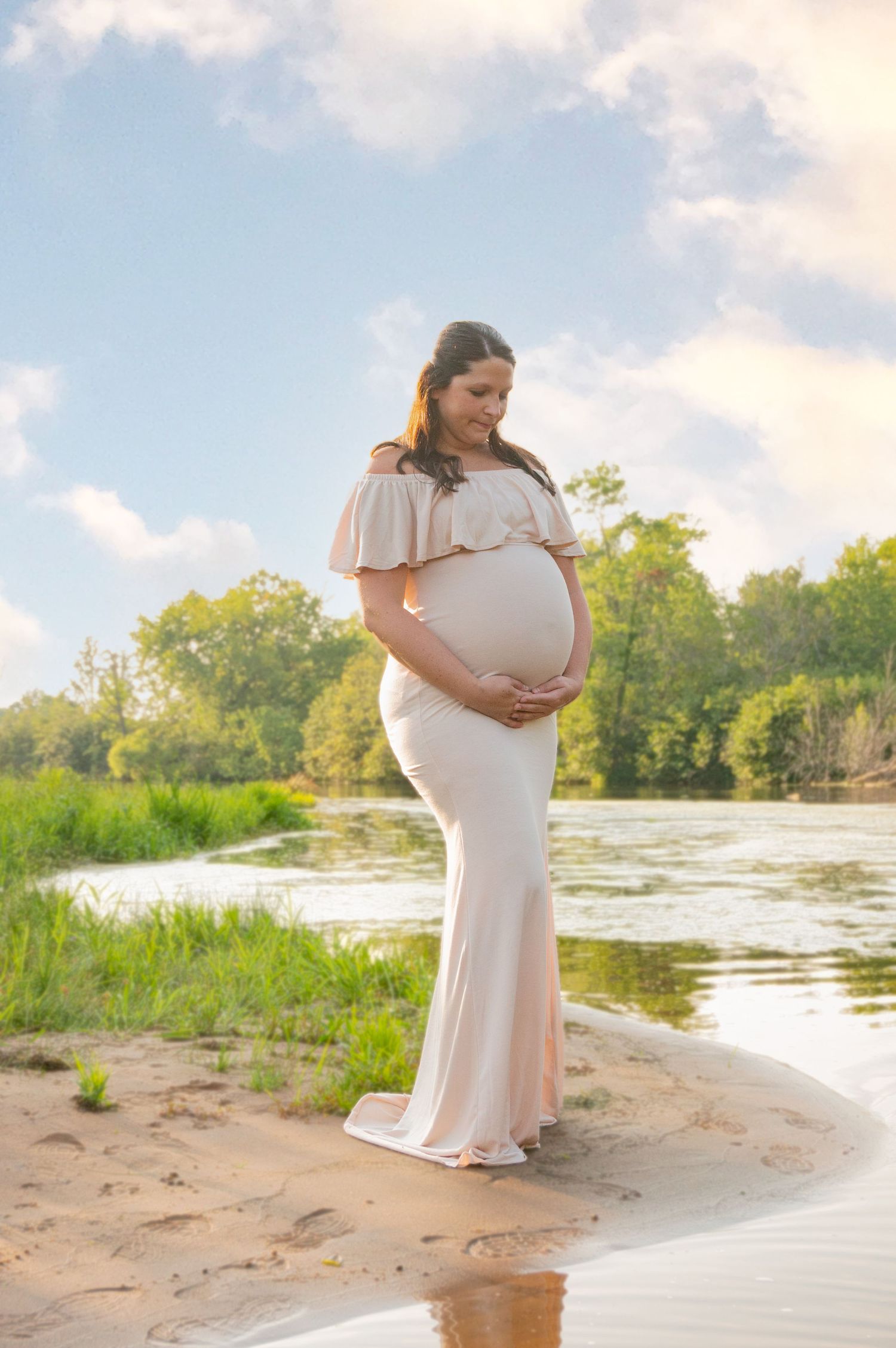 Maternity photoshoot in a flowing beige dress at sunset by a serene waterfront.
