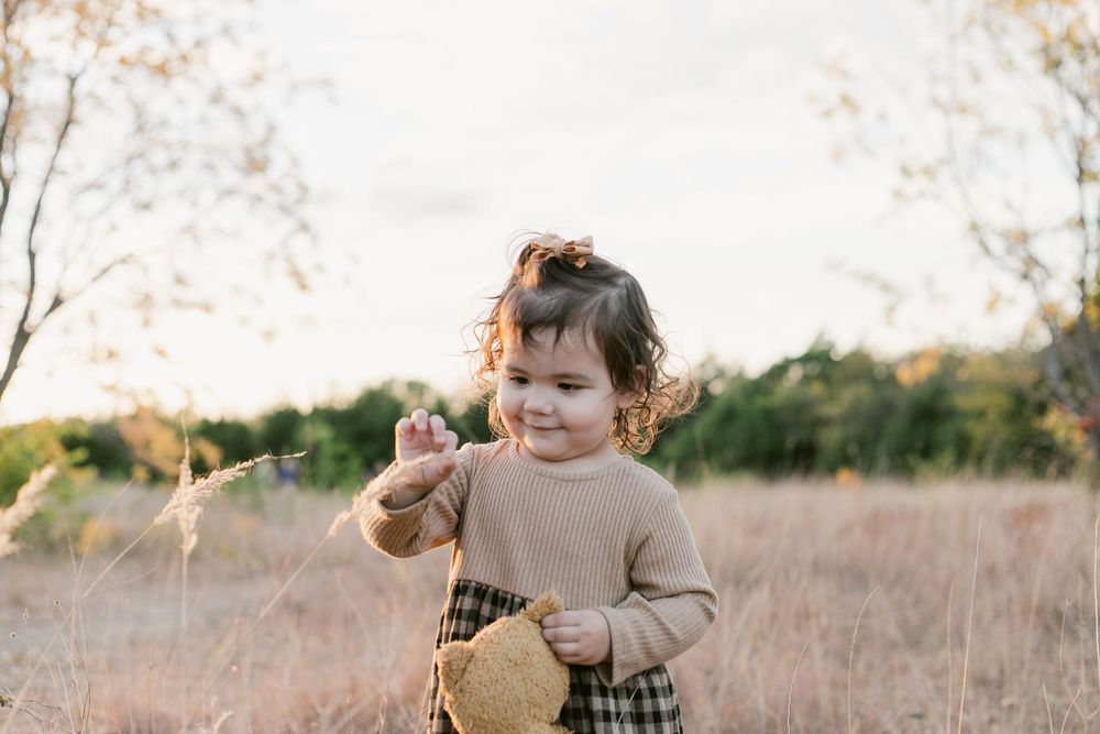 Candid outdoor portrait of a toddler girl holding a stuffed animal while playing in a grassy golden-hour field.