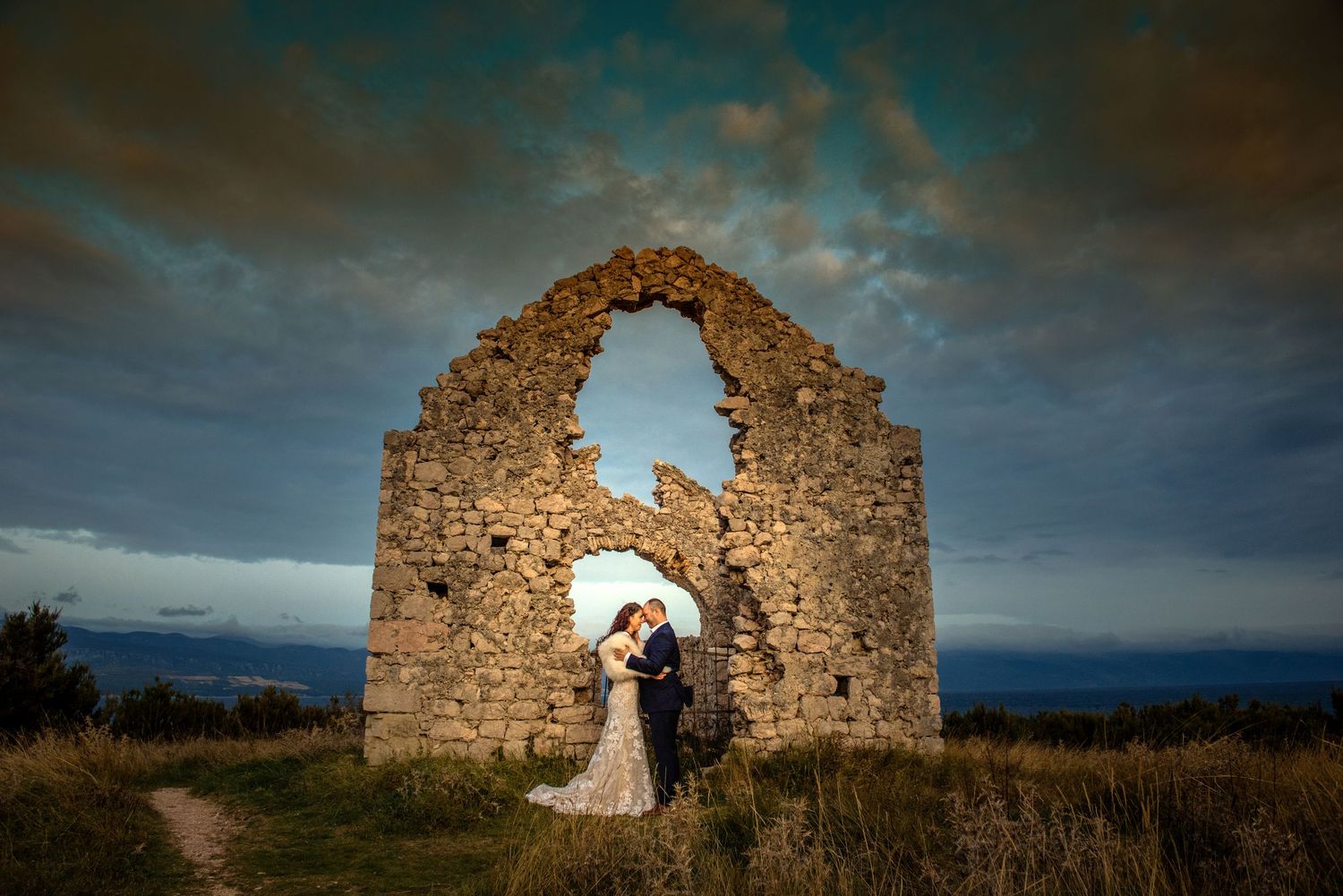 Dramatic wedding photo featuring stone ruins arch against moody sky at sunset.