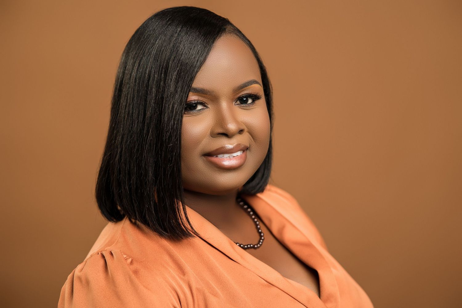 Professional Black woman in orange blouse with straight hair against warm brown background.