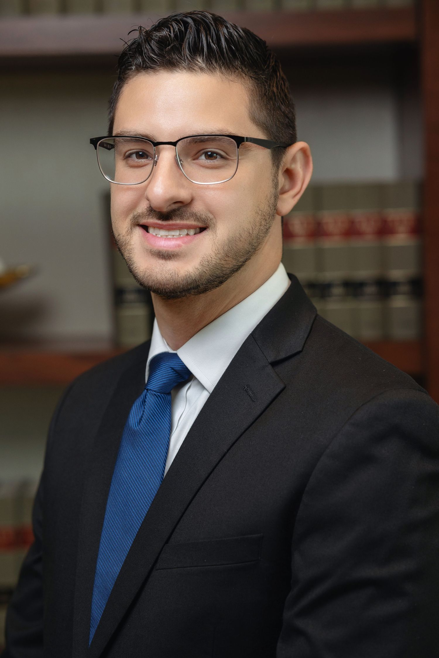 Professional headshot of a young man in a dark suit and blue tie with glasses in front of law books.