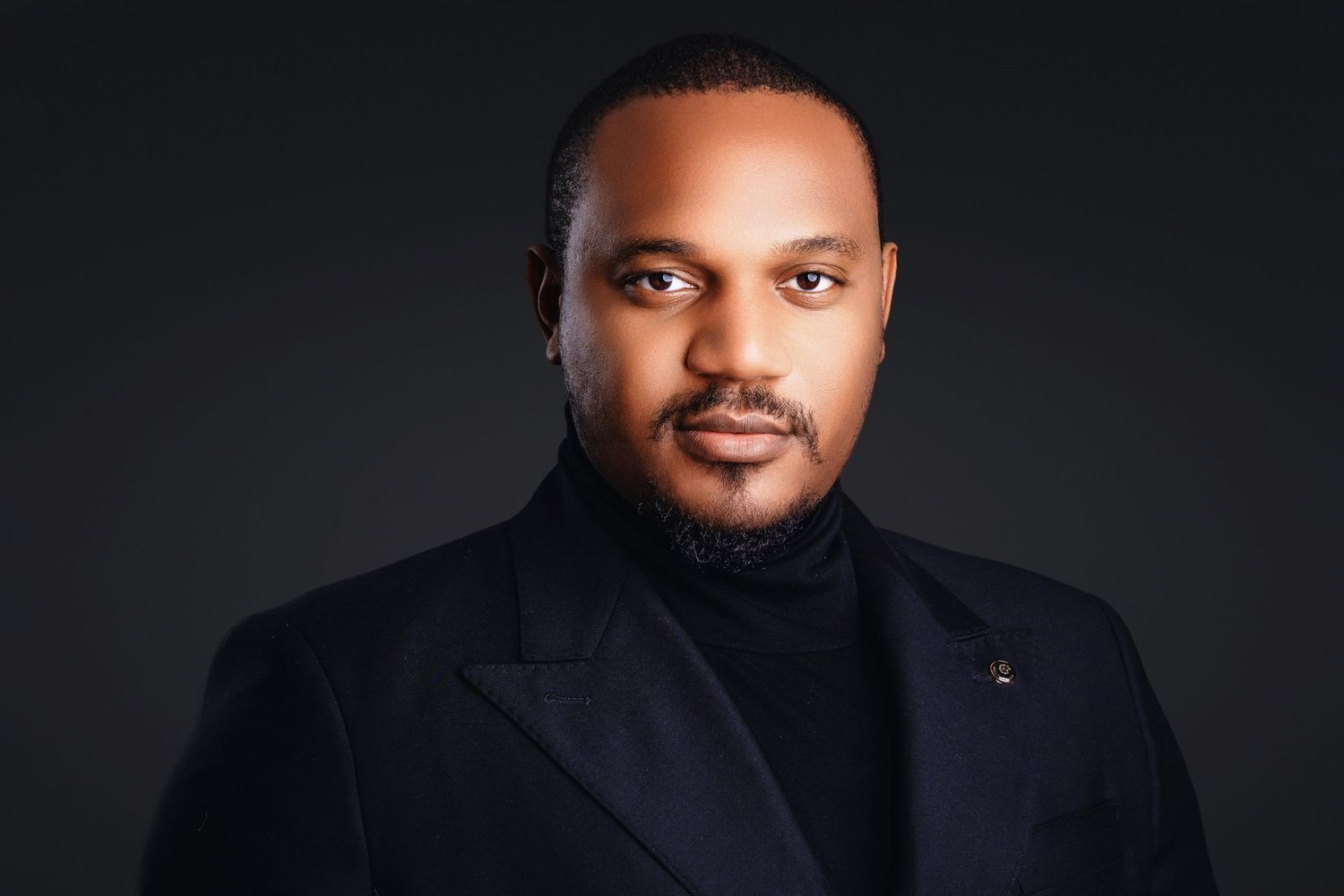 Confident man in black turtleneck coat posing for professional portrait on dark background.
