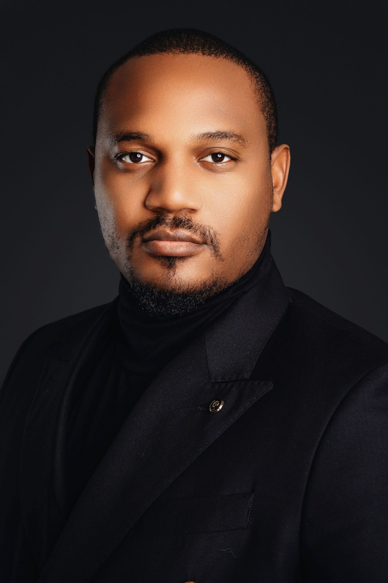 Professional headshot of man in black blazer and turtleneck against dark studio background.