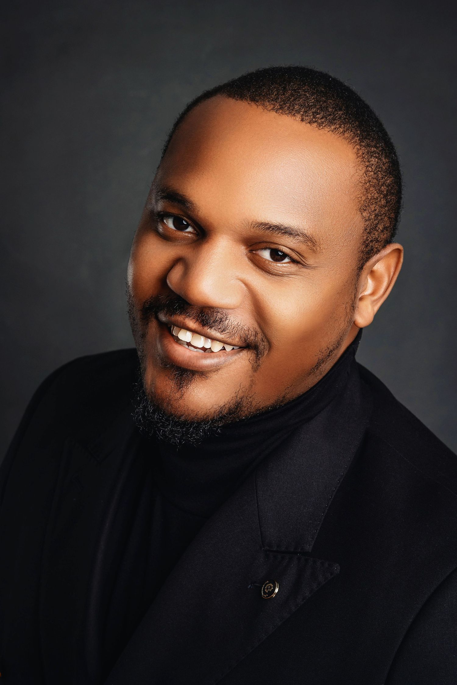 Smiling man in black turtleneck and blazer posing for professional headshot on dark background.