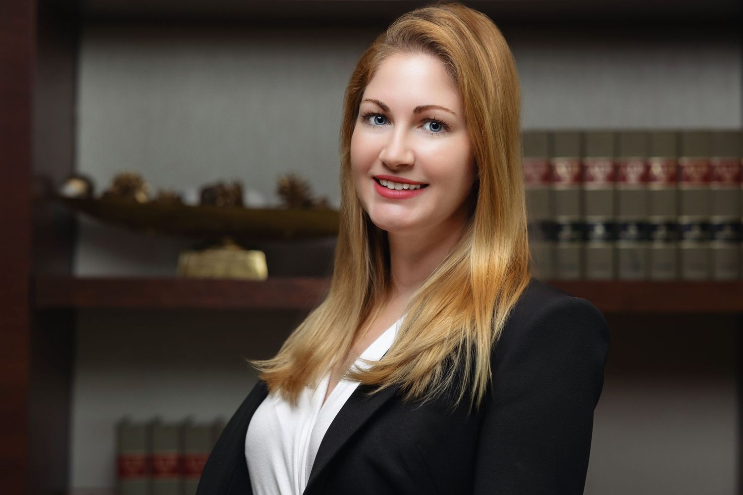 Professional blonde woman in black blazer smiling before law office bookshelf.