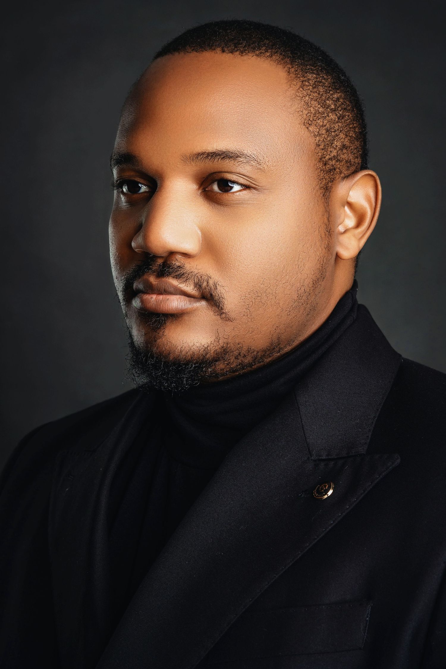 Man in black turtleneck and blazer with serious expression in professional studio headshot.