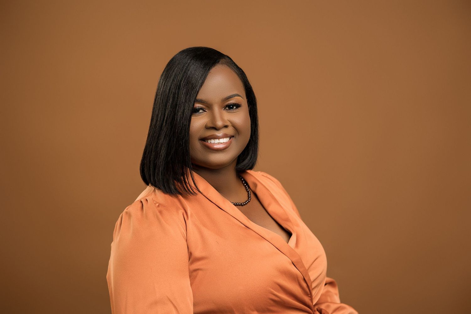 Smiling Black woman in orange top with straight bob haircut on brown background.