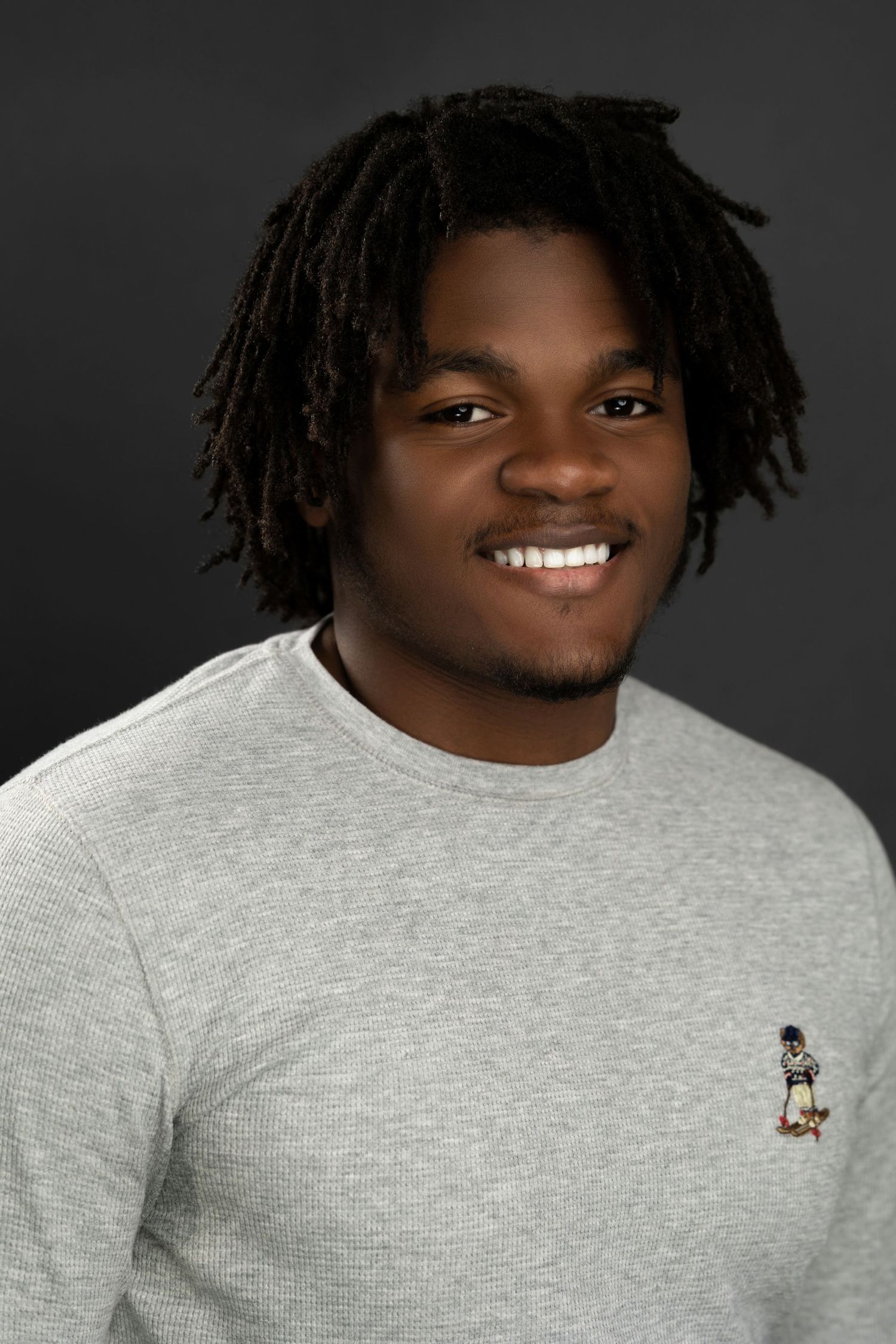 Smiling young man with dreadlocks in gray sweater against dark background.