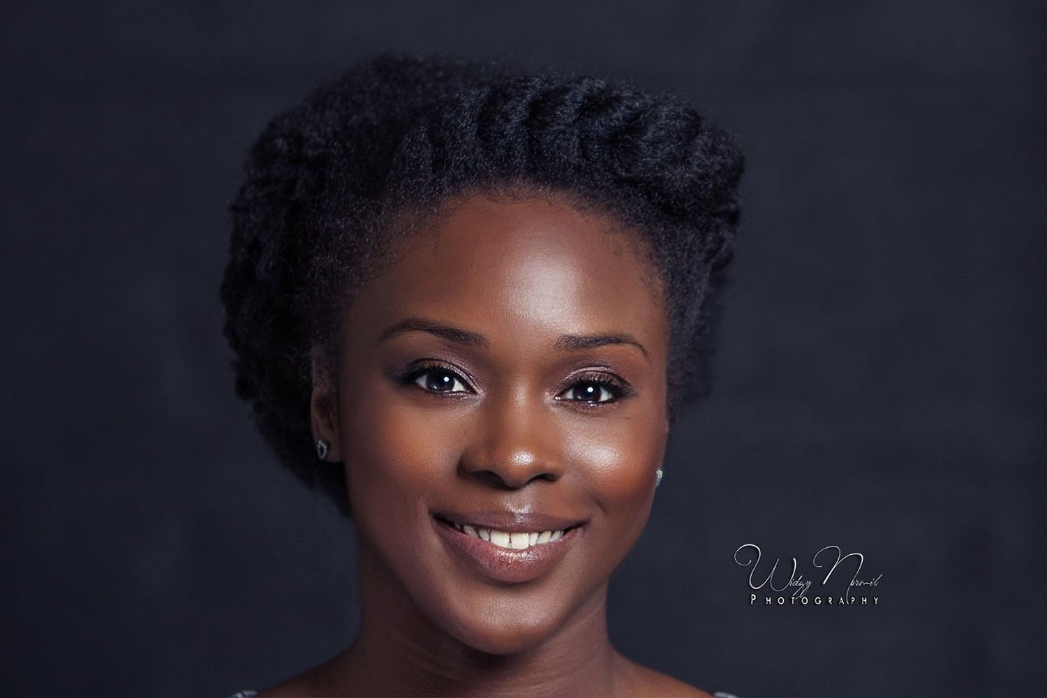 Young Black woman with elegant twisted updo hairstyle smiling warmly against dark studio background.