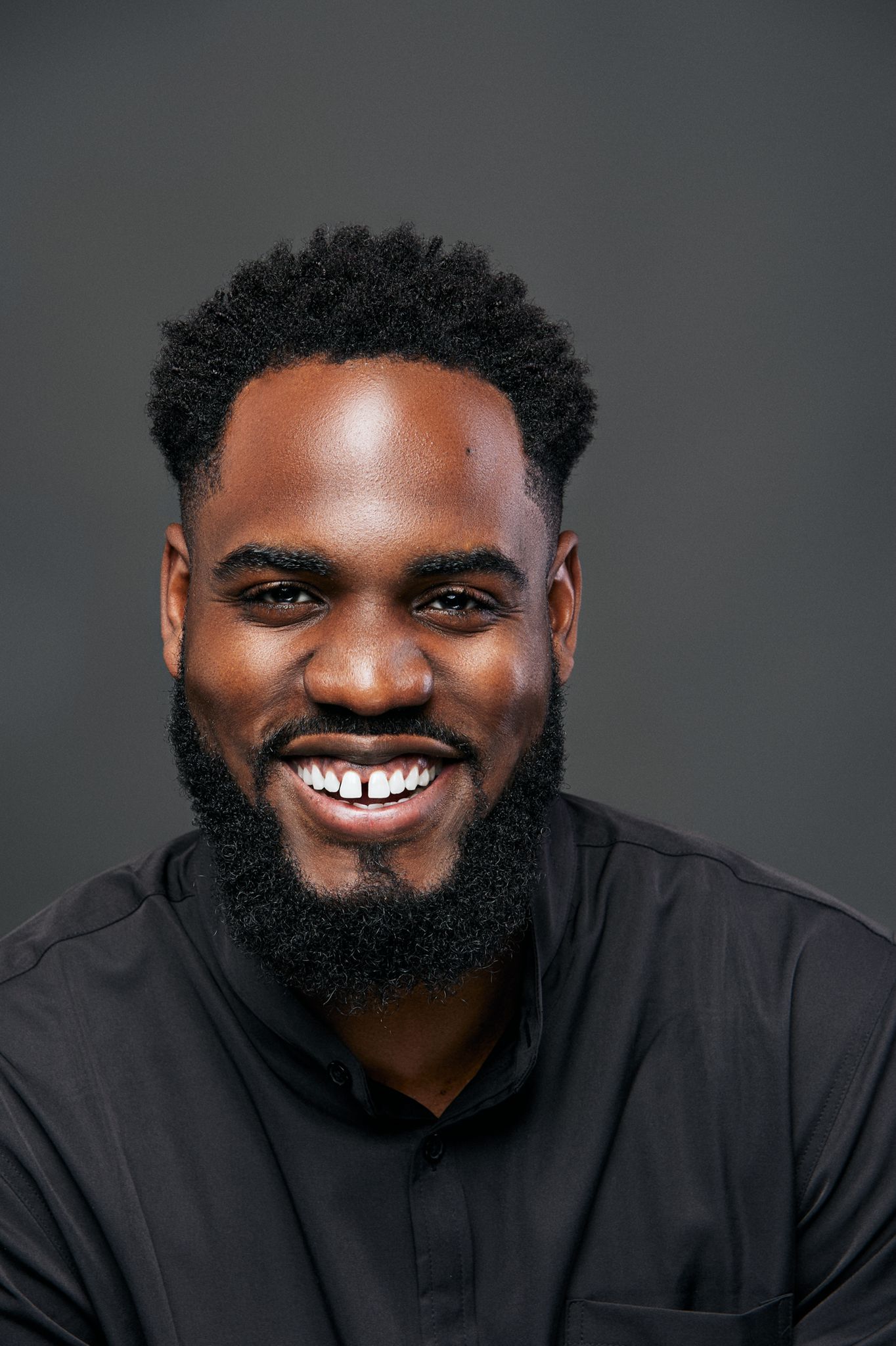 Smiling Black man with well-groomed beard wearing black shirt against gray studio background.
