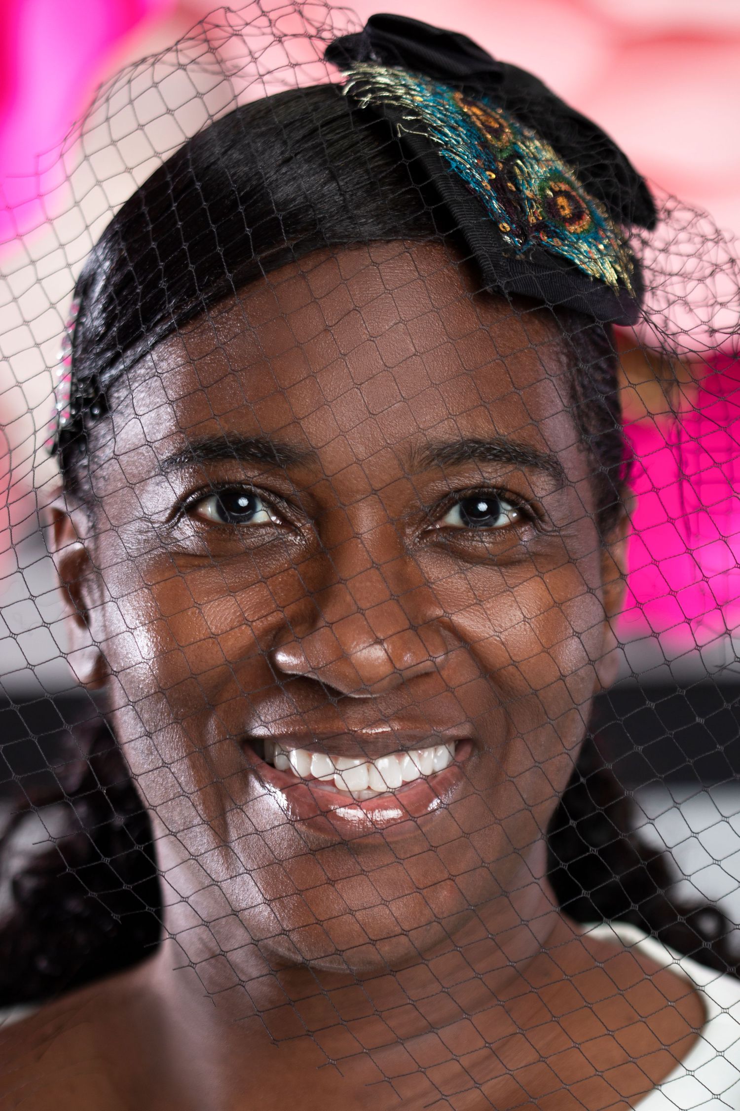 Close-up portrait of smiling woman wearing black birdcage veil fascinator with peacock feather embellishment.