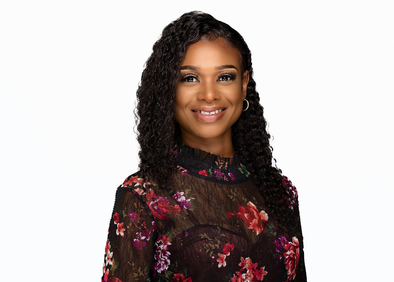 Young woman with curly hair in floral blouse smiles for professional headshot on white background.