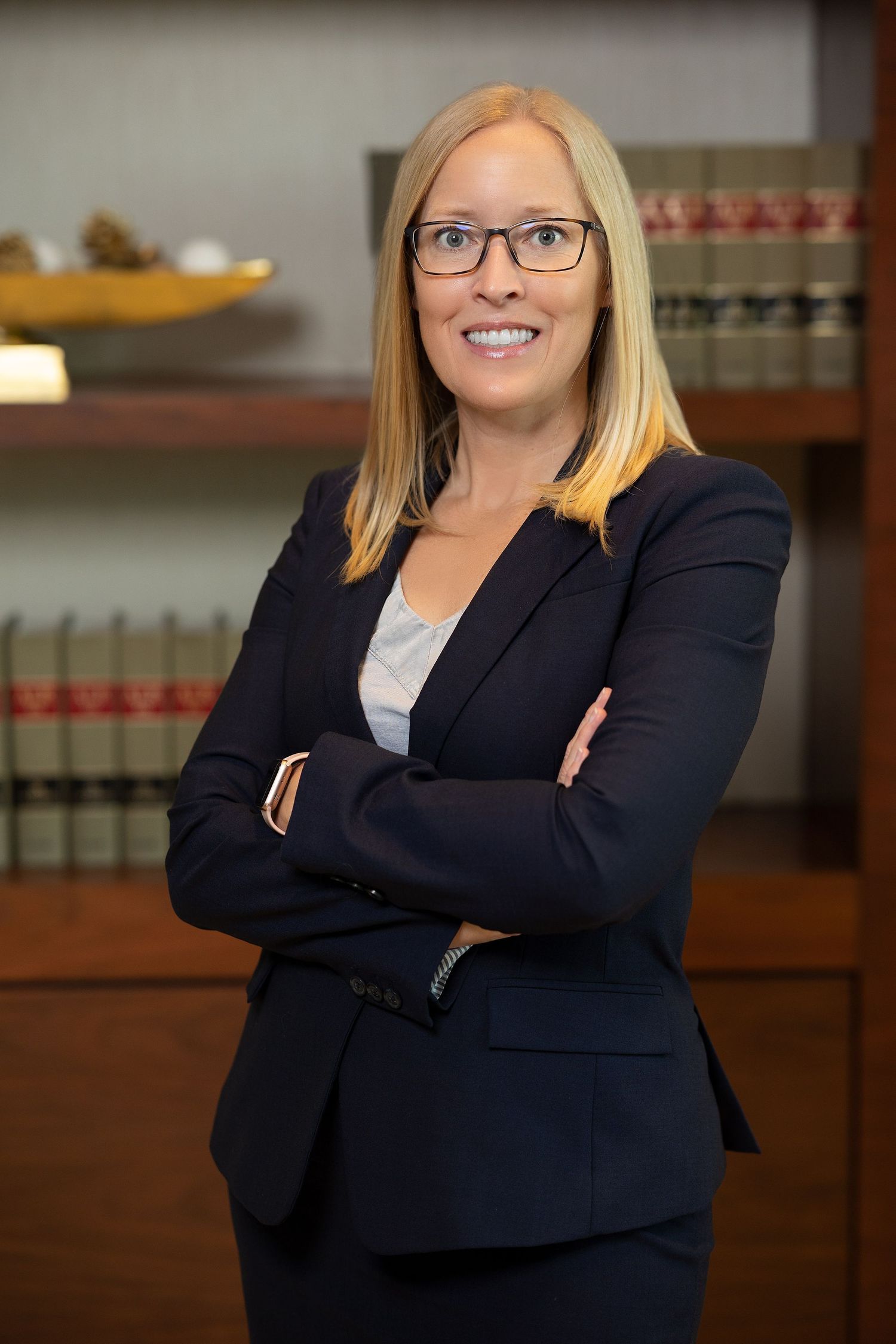 Blonde woman in navy blazer with arms crossed smiles for professional headshot in law office setting.