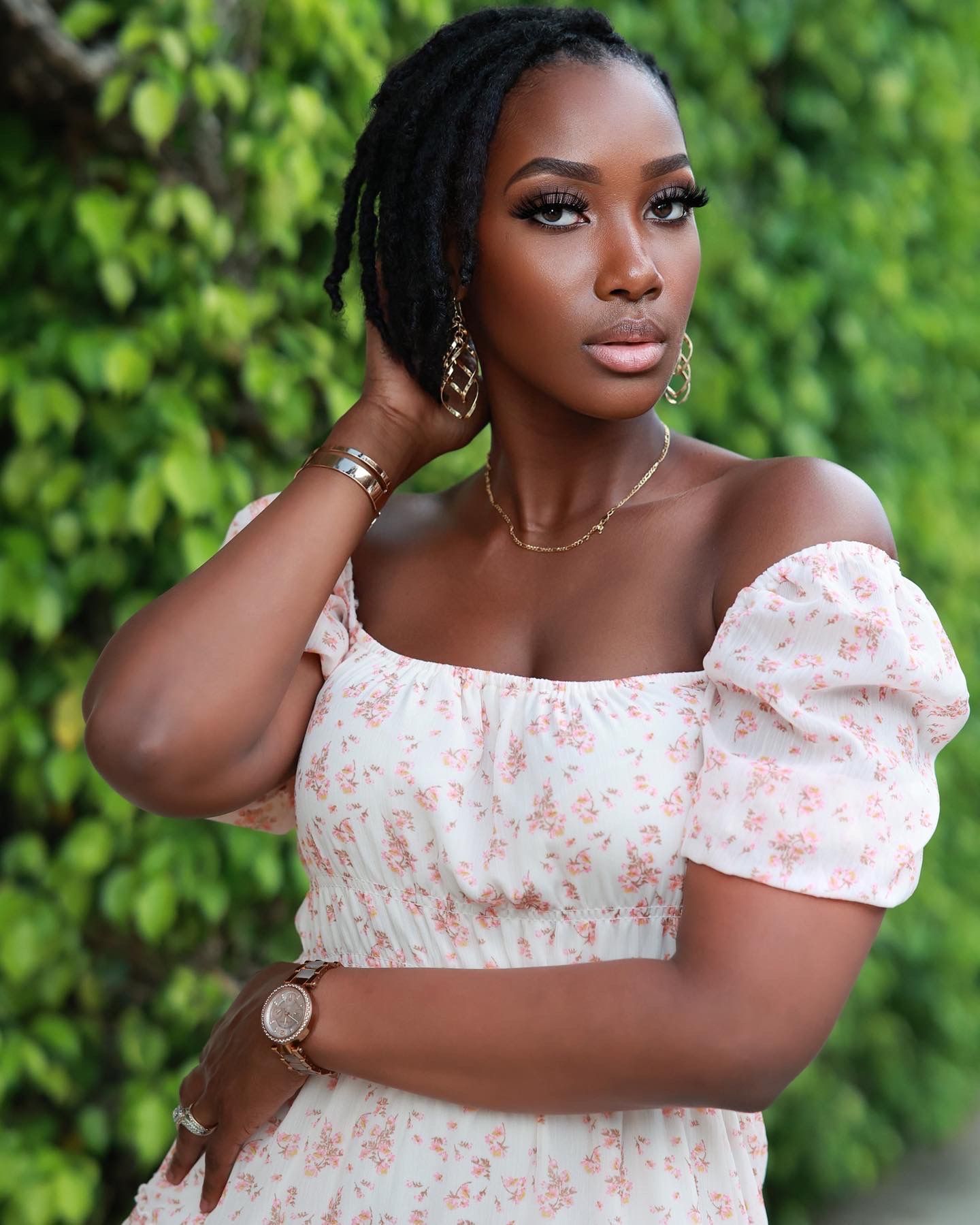 Elegant Black woman in off-shoulder floral dress poses outdoors with lush green foliage background.
