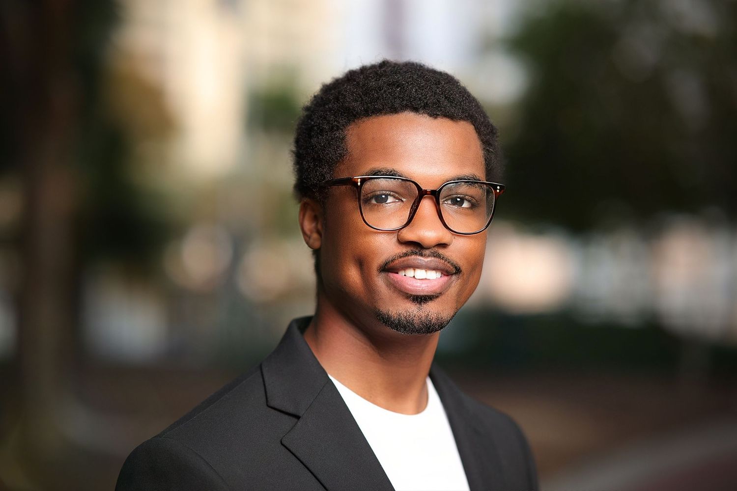 Young man in glasses and blazer smiling in an outdoor urban portrait with bokeh background.