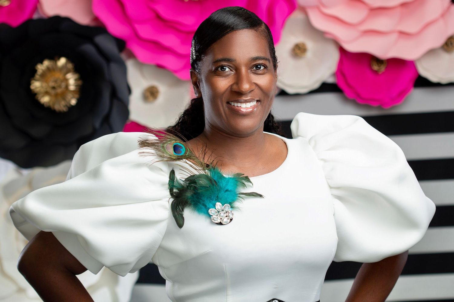 Black woman in white puffed-sleeve dress with peacock feather brooch smiling by paper flower wall.