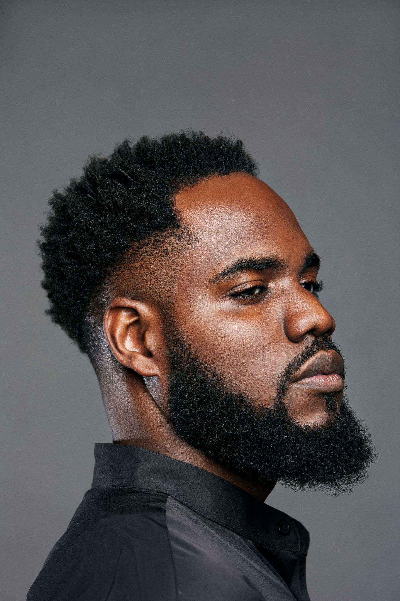 Side profile of Black man with stylish beard and fade haircut wearing black shirt on gray background.