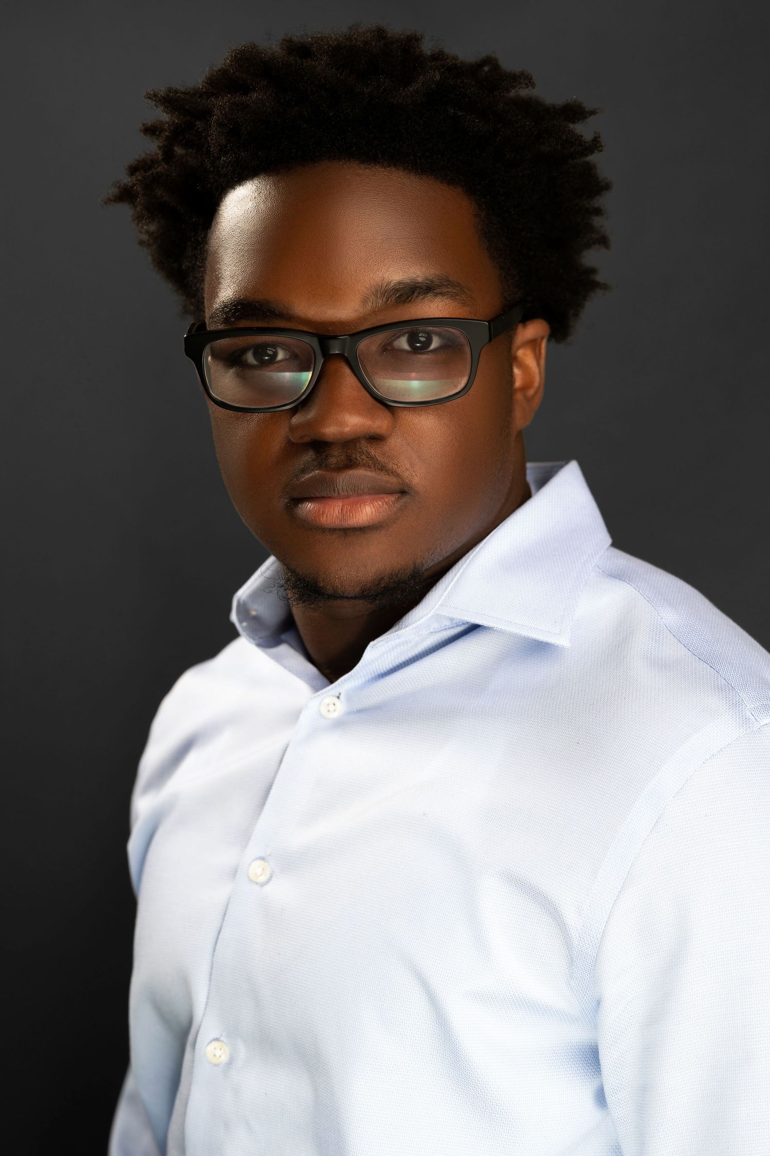 Young Black man in light blue dress shirt and black glasses poses for professional headshot against dark background.