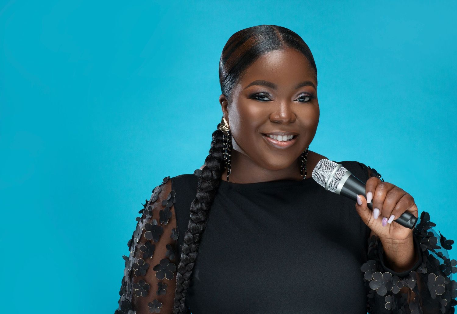 Cheerful Black woman with braids holding microphone laughing in black lace top on teal background.