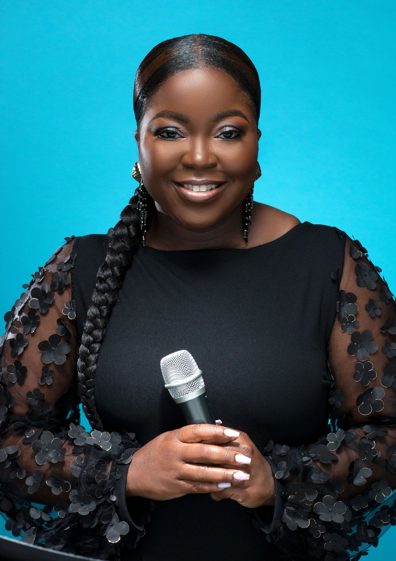 Joyful Black woman holding microphone in black floral lace top smiling against teal background.