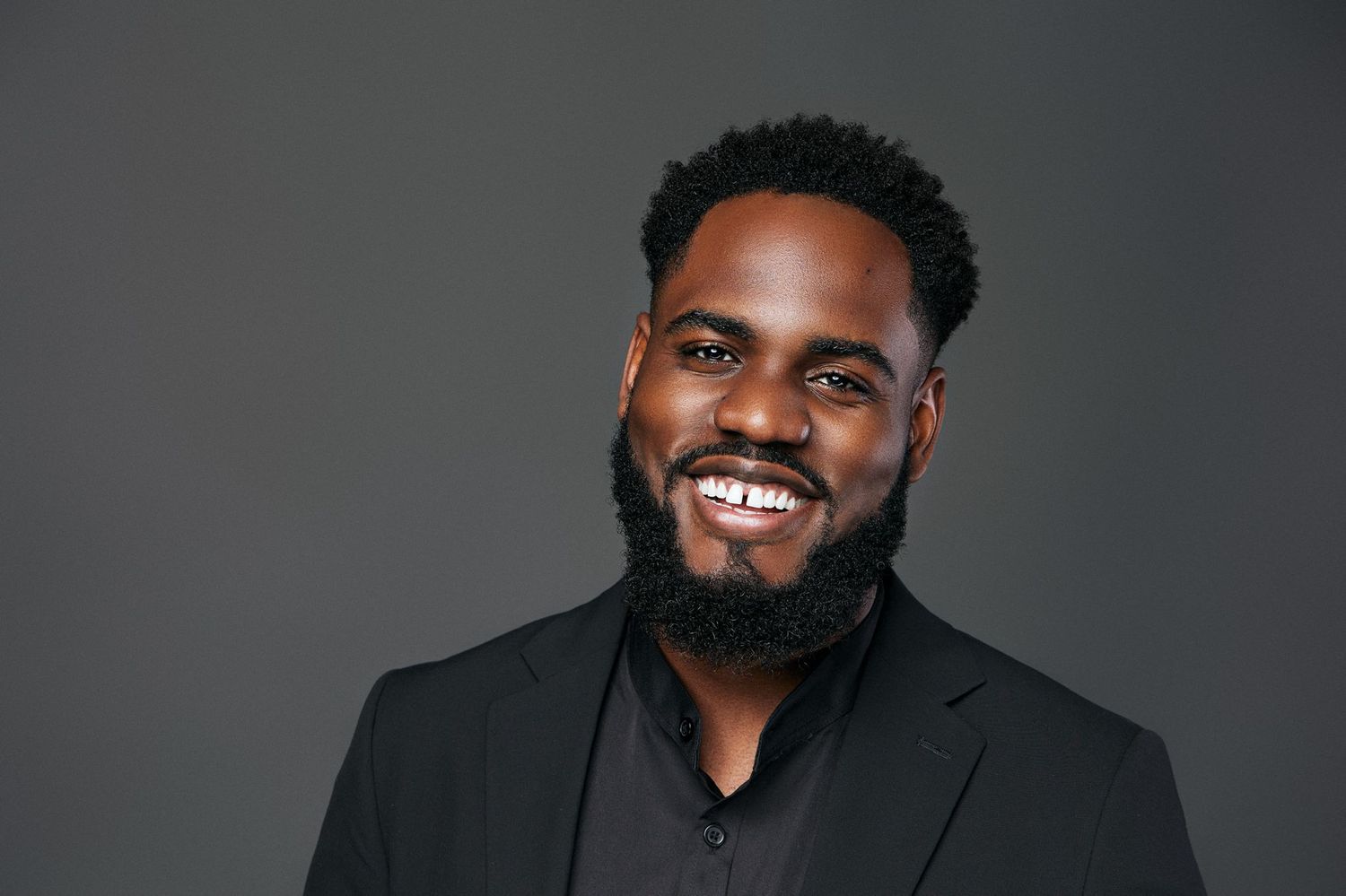 Confident Black man with beard smiling broadly in black blazer against gray studio background.