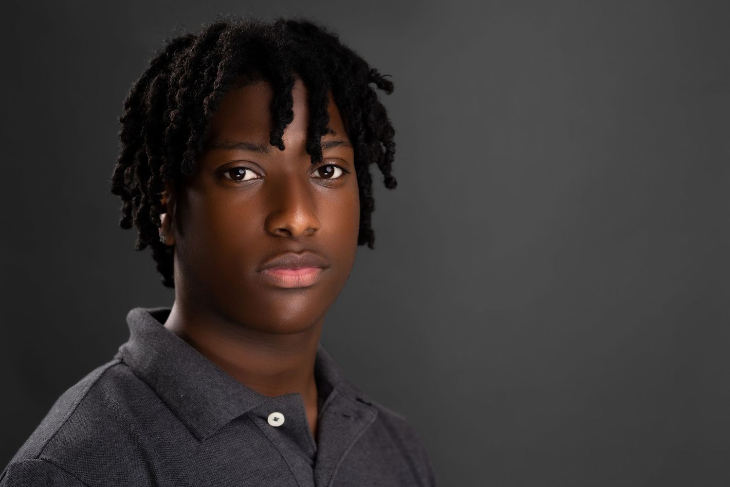 Serious young man with dreadlocks wearing gray polo shirt against dark background.