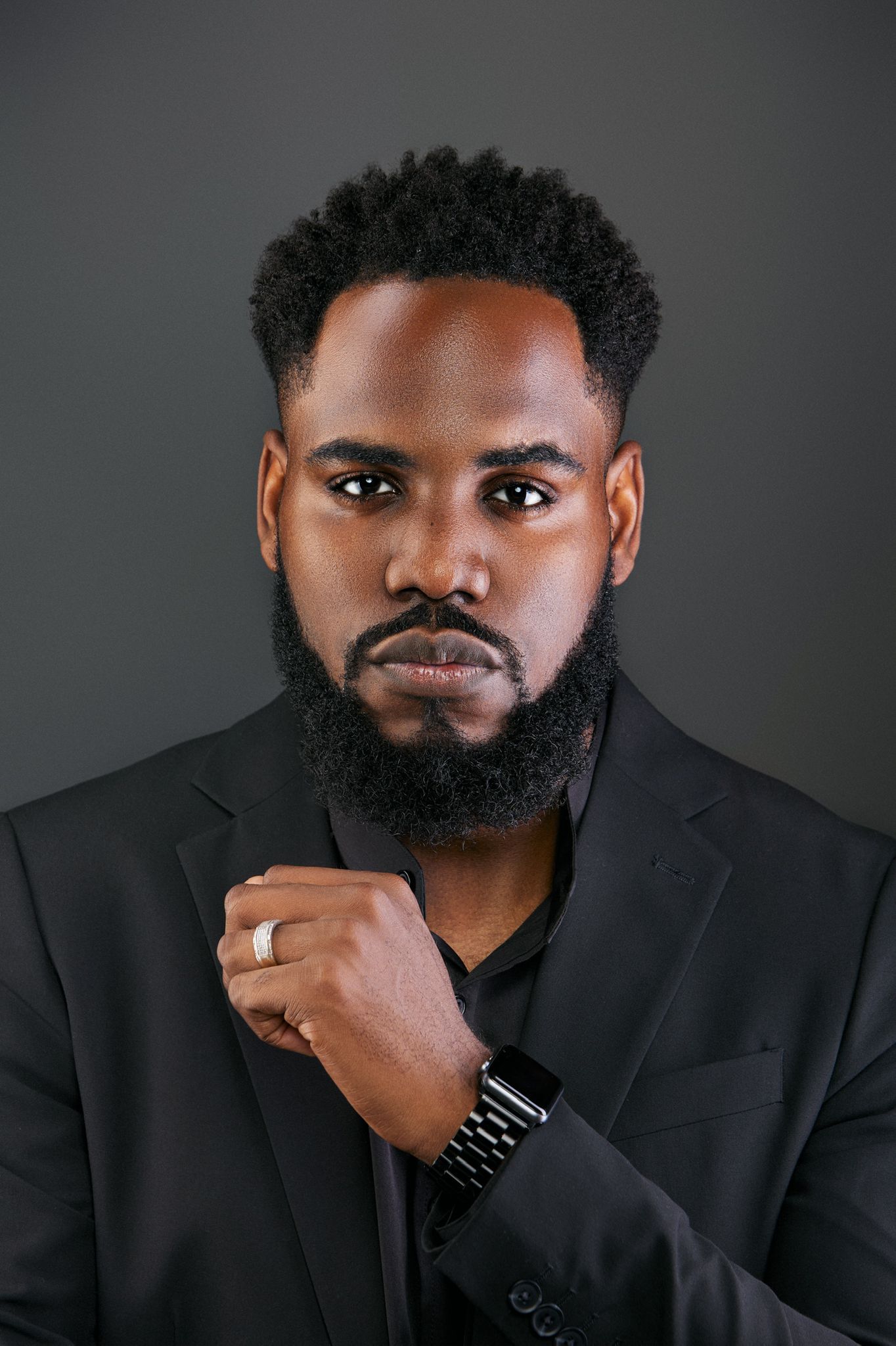 Serious Black man with beard in black suit holding lapel with watch and ring accessory.
