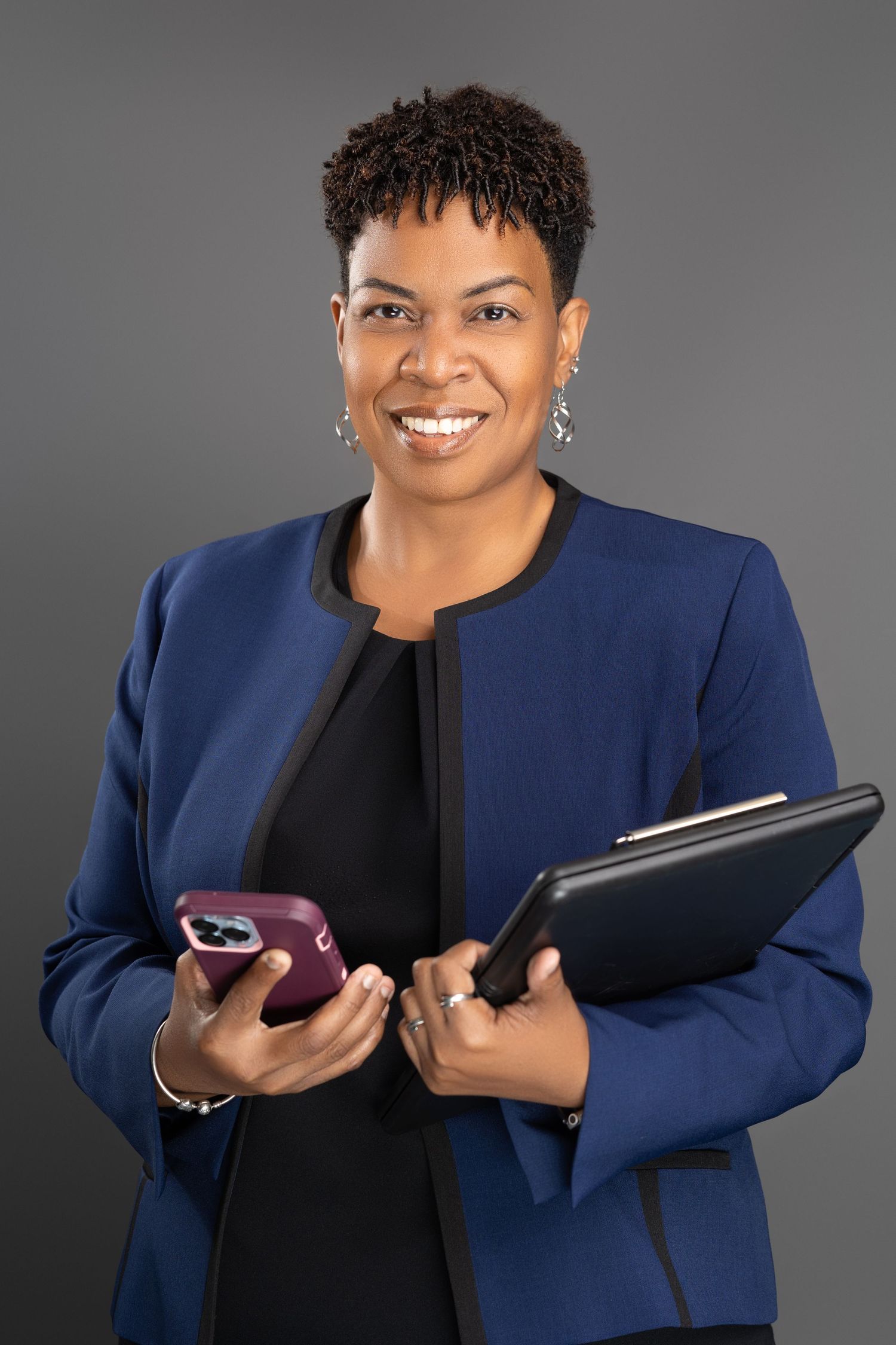 Professional Black woman in blue blazer holds phone and portfolio while smiling for business portrait.