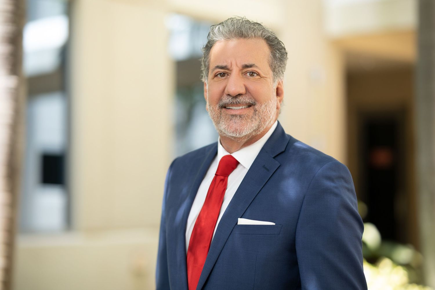 Smiling middle-aged man in navy suit and red tie poses for professional business portrait in bright lobby.