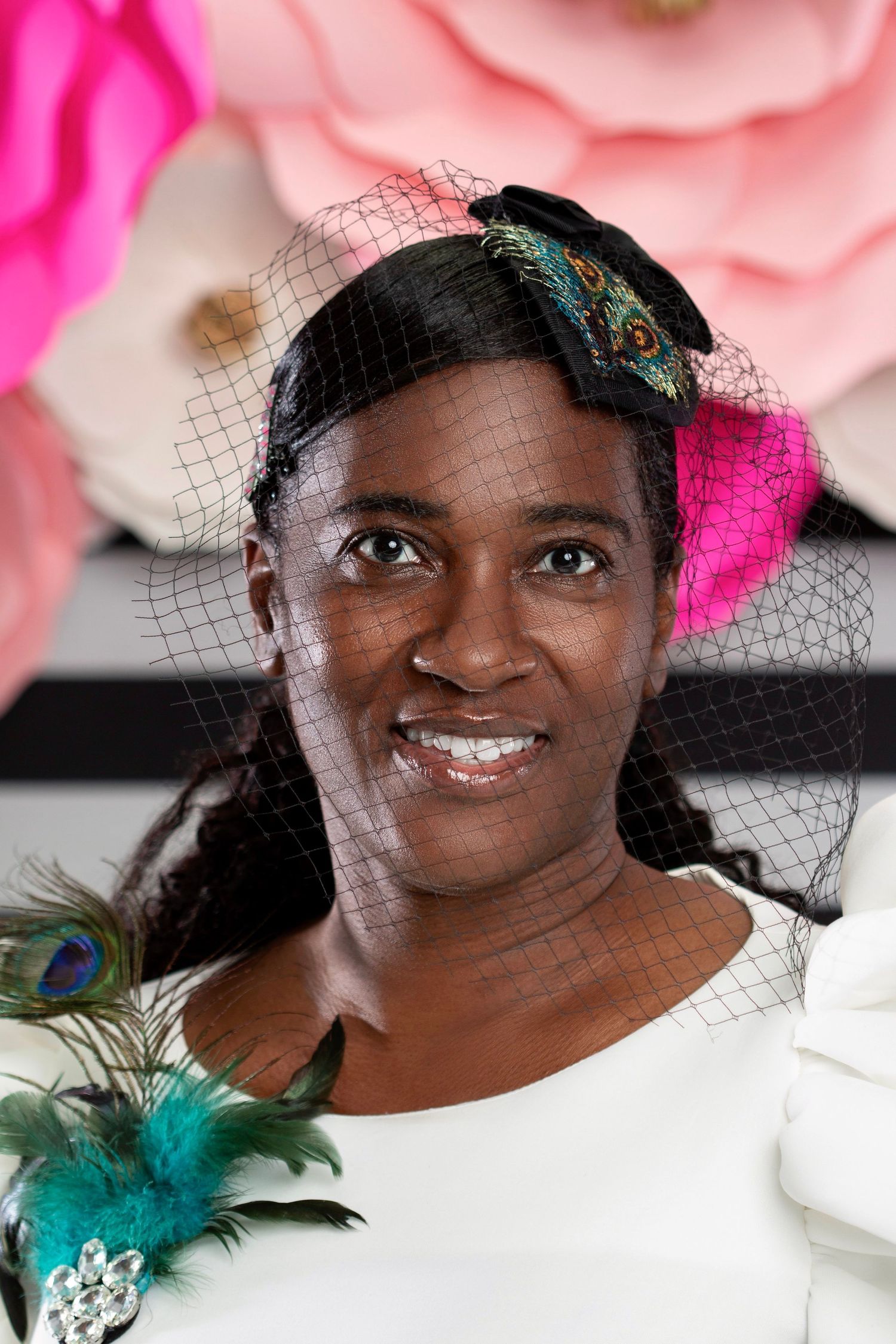 Woman smiling in elegant black fascinator hat with peacock feather details and birdcage veil, surrounded by paper flowers.