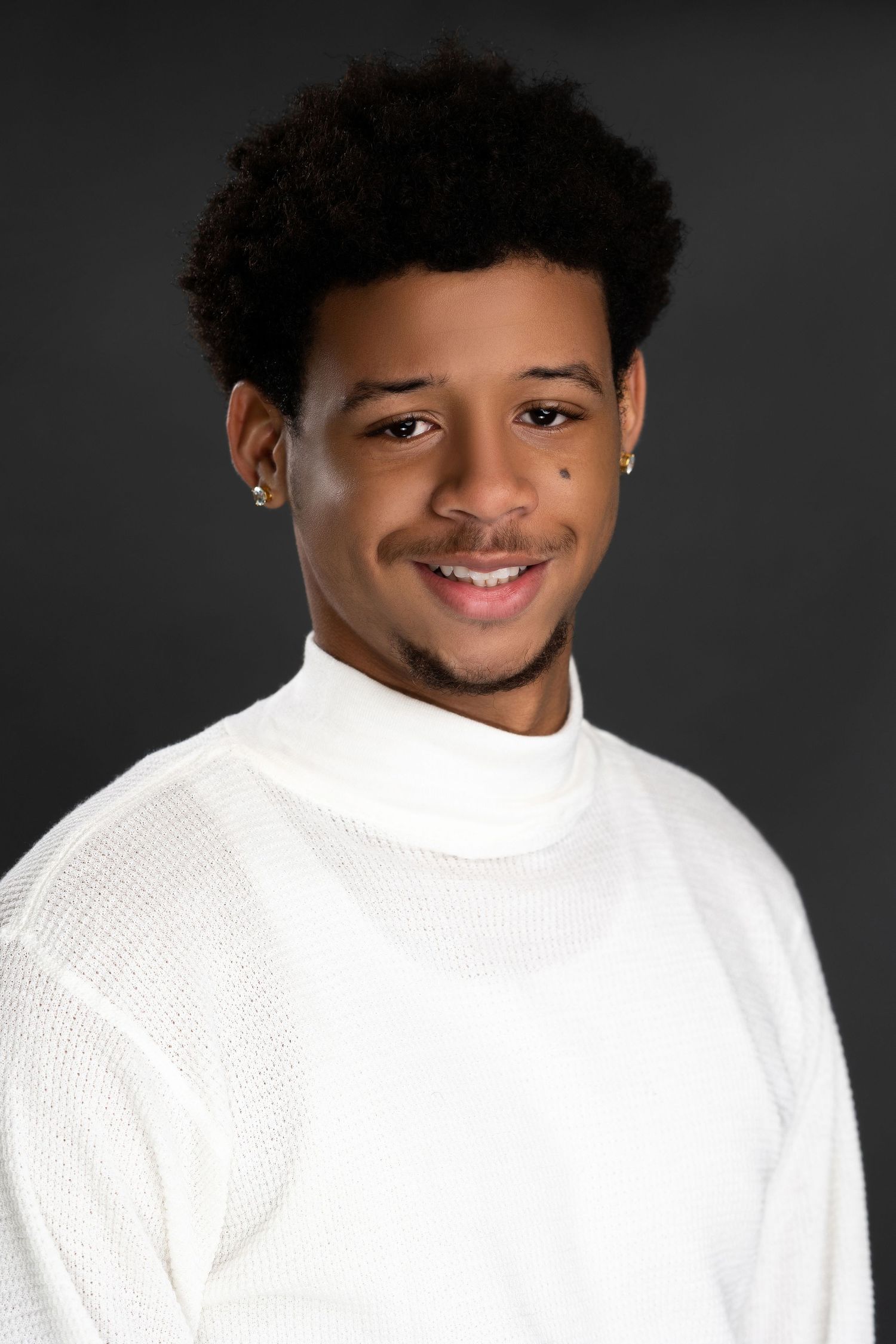 Smiling young man in white turtleneck sweater posing against dark background.