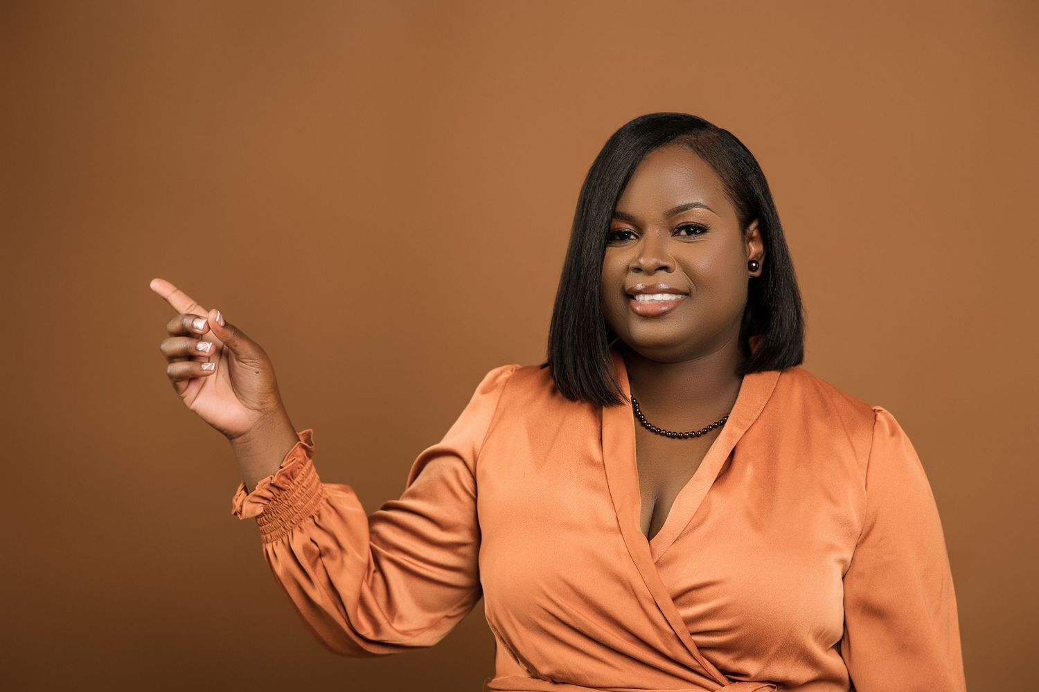 Smiling Black woman in orange satin wrap blouse pointing sideways against brown background.