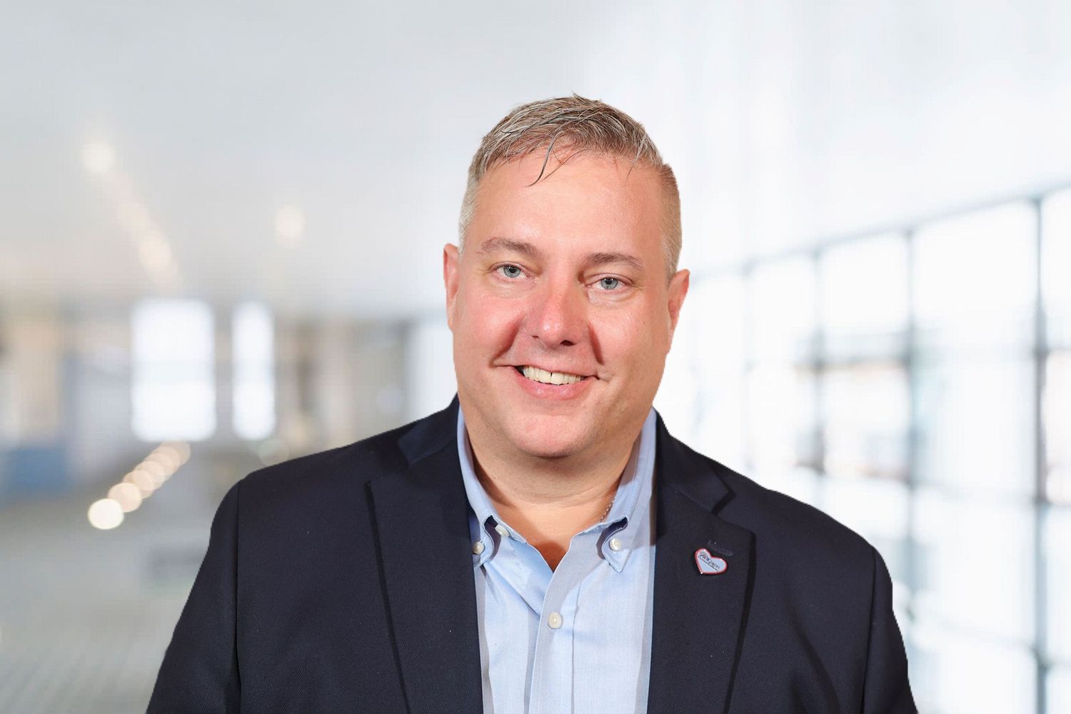Smiling man in blazer with lapel pin posing in a modern bright hallway.